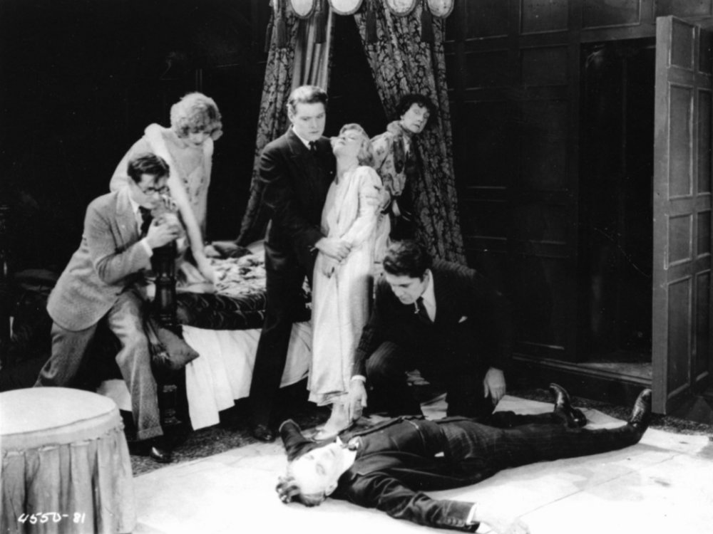 Eine Gruppe Menschen in Kleidung der 1920er schaut auf einen am Boden liegenden, dem Anschein nach ermordeten, Mann