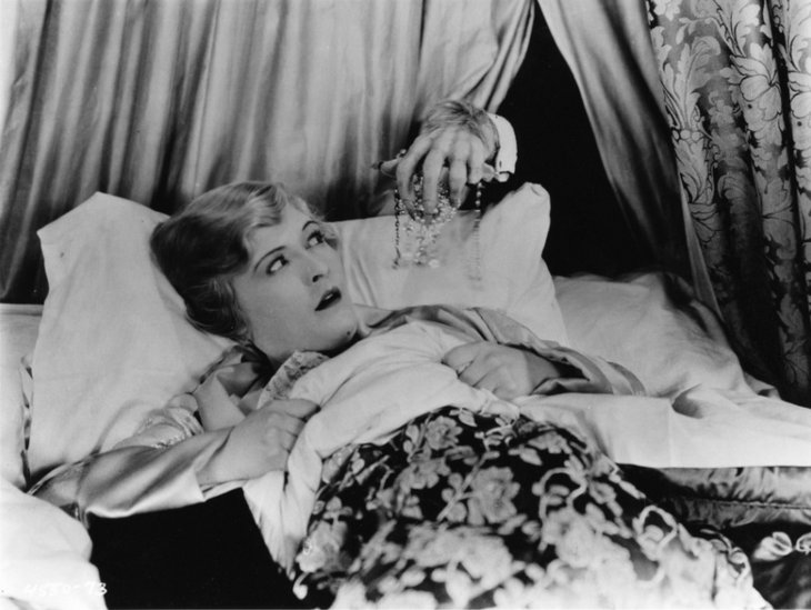 Eine blonde Frau liegt im Bett und guckt ängstlich. Von hinten greift die haarige Hand eines Monsters nach ihr