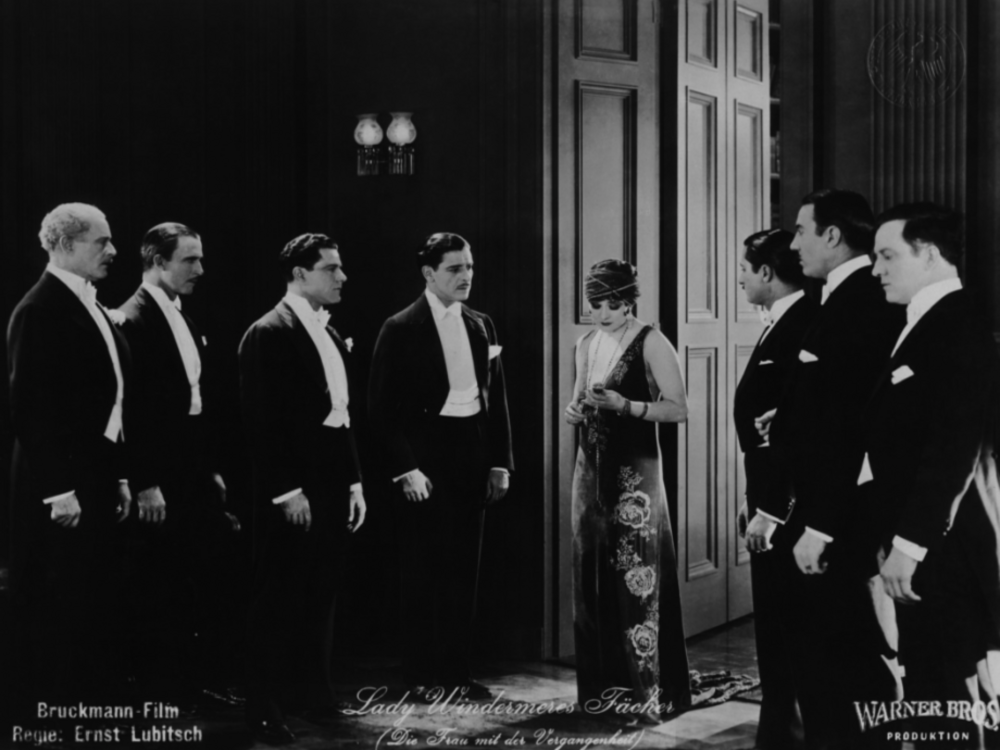 Eine dunkelhaarige Frau im langen dunklen 1920-er Abendkleid steht inmitten zwei Reihen Männern in Smokings