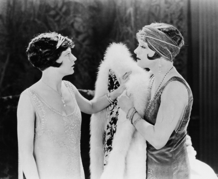 Zwei dunkelhaarige Frauen mit Frisuren und Kleidung im 1920er-Jahre-Style stehen sich gegenüber. Eine der Frauen hält einen weißen Pelz. 