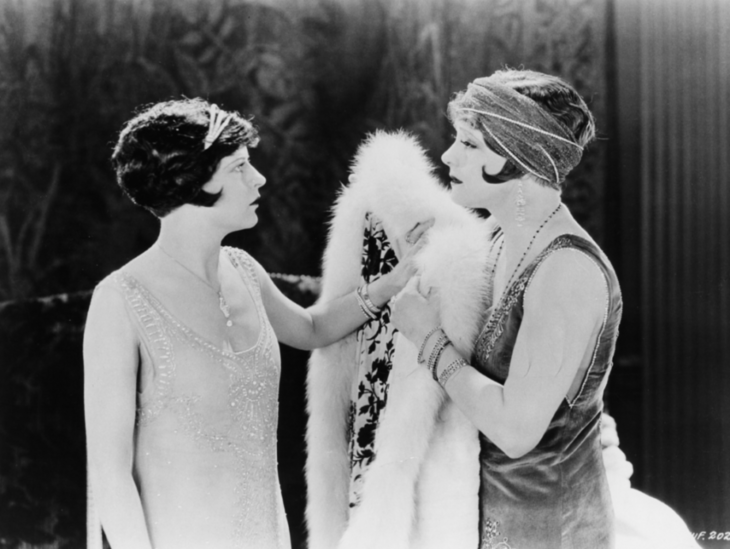 Zwei dunkelhaarige Frauen mit Frisuren und Kleidung im 1920er-Jahre-Style stehen sich gegenüber. Eine der Frauen hält einen weißen Pelz. 